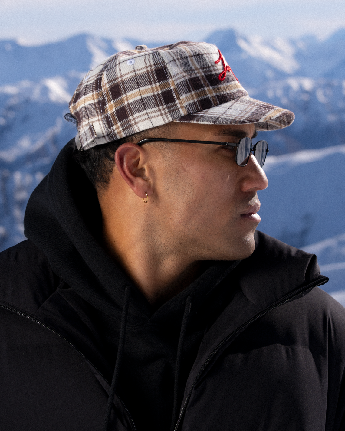 A man in a black winter jacket and sunglasses wearing a brown, beige, and white plaid baseball cap with a red logo, set against a snowy mountain background.